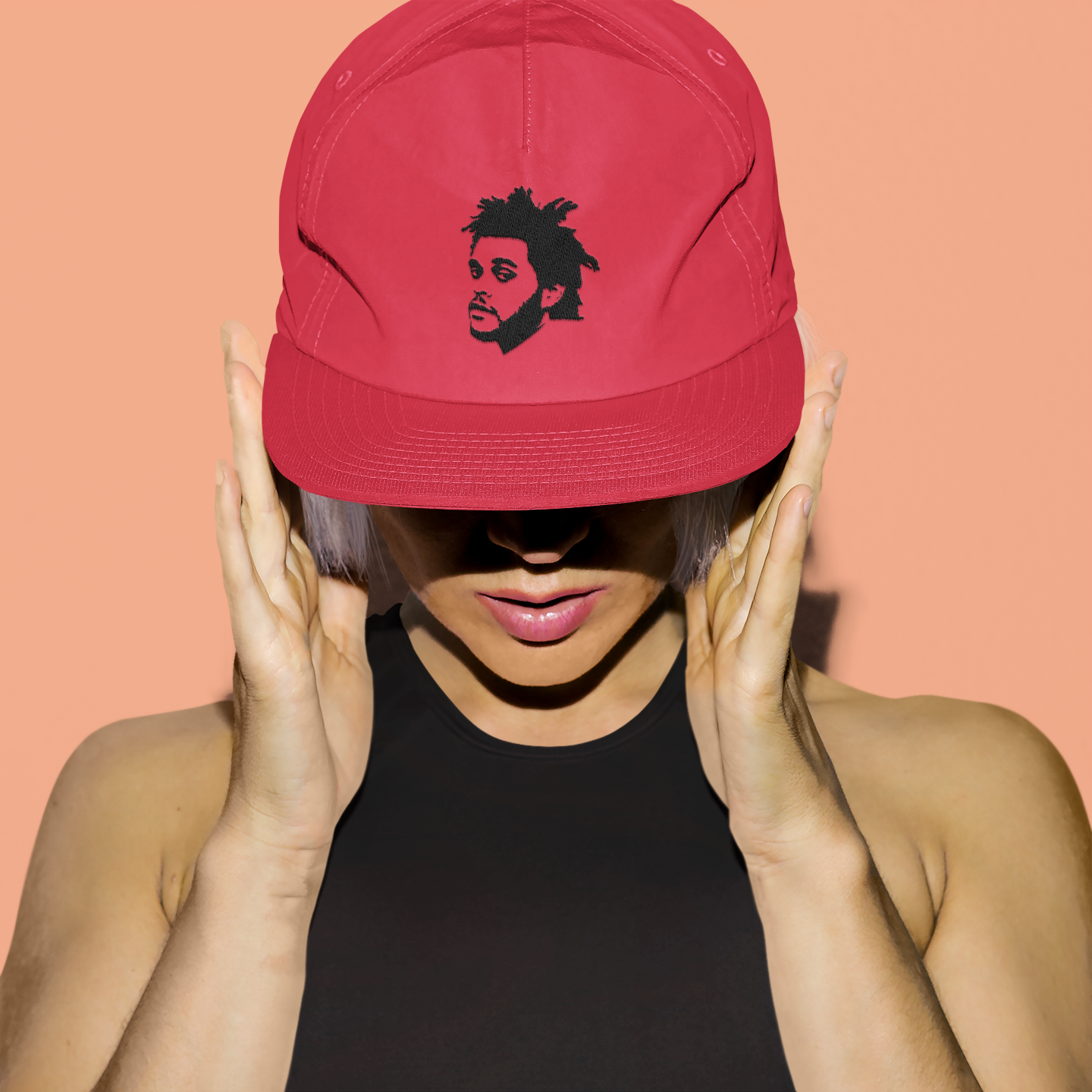 Woman wearing a red snapback hat embroidered with The Weeknd’s face, styled with a black crop top against a peach background – AmHype USA concert merch.