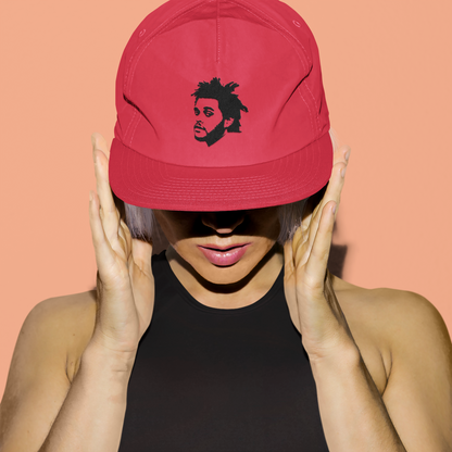 Woman wearing a red snapback hat embroidered with The Weeknd’s face, styled with a black crop top against a peach background – AmHype USA concert merch.